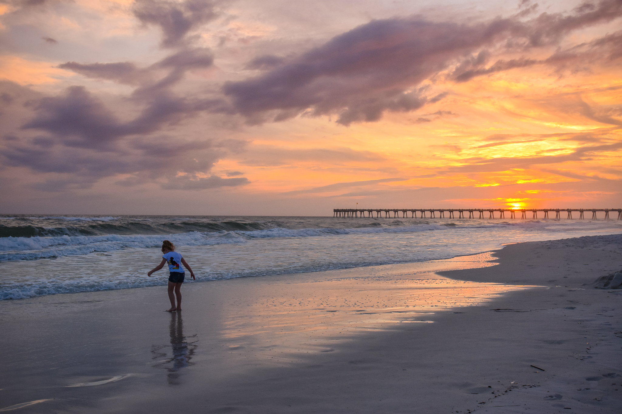 The sun setting behind the Pensacola pier with a young girl playing in the surf